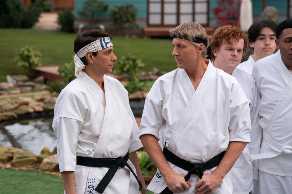 Ralph Macchio as Daniel LaRusso and William Zabka as Johnny Lawrence stand face-to-face in martial arts uniforms, with younger students standing behind them in a peaceful dojo setting with a small pond and garden.