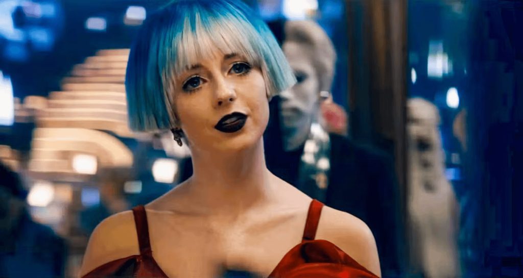 Rachel Garrett, played by Kacey Rohl, with a striking blue and silver bob haircut, wearing dark lipstick and a red dress, gazes confidently at the camera in a futuristic setting, surrounded by blurred lights and figures.