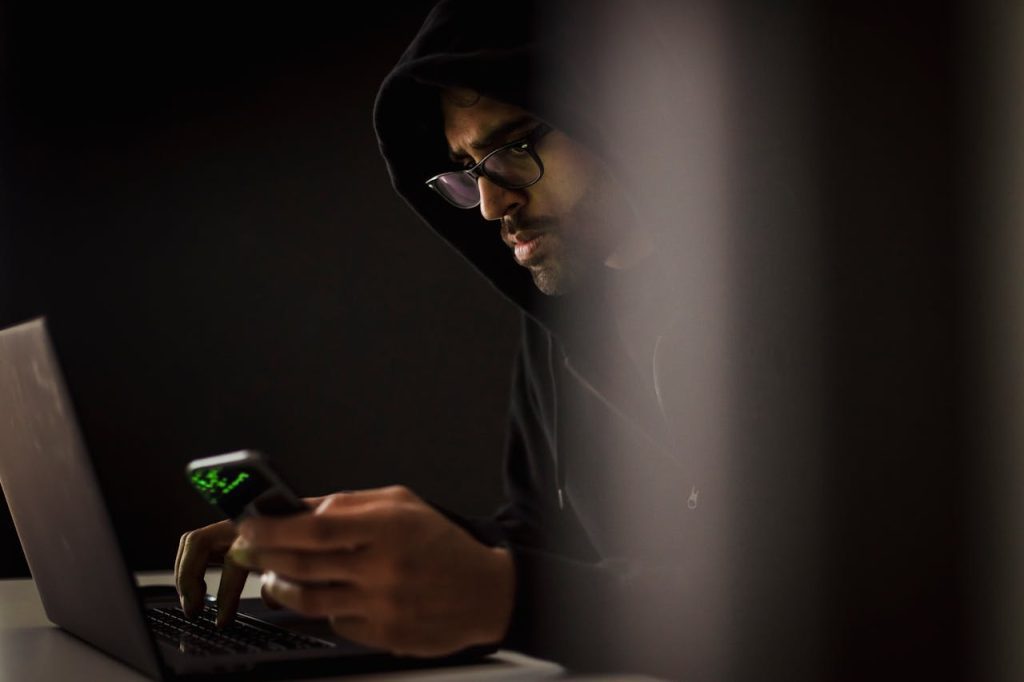 Phone tracking privacy A man wearing glasses and a hoodie focuses on his smartphone while typing on a laptop in a dark room.